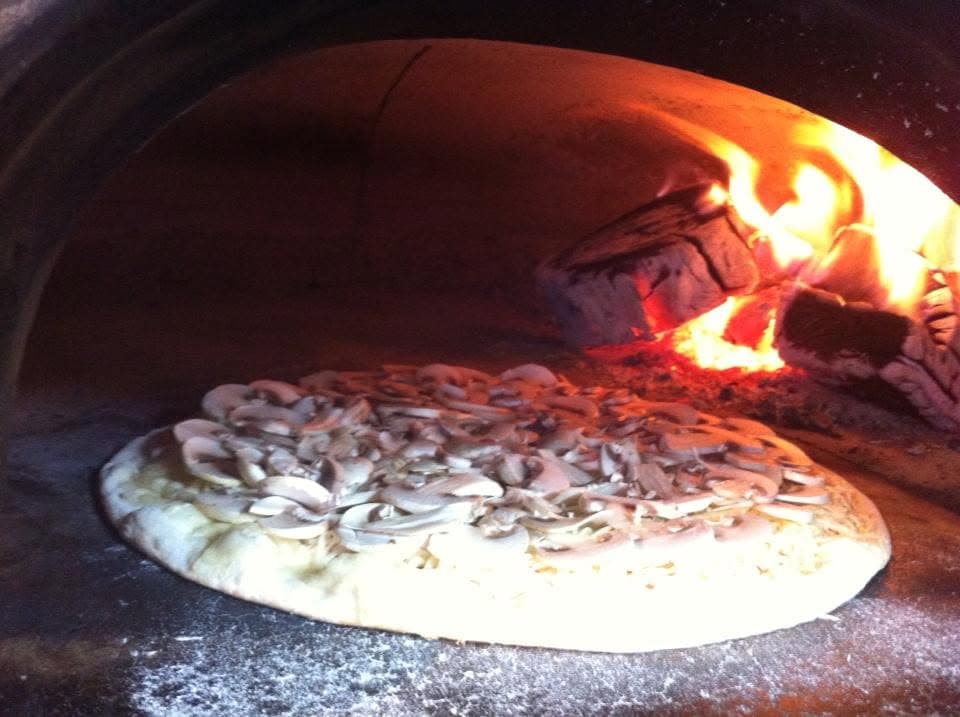 Une pizza aux champignons dans le four à bois, les bûches en flammes juste à côté